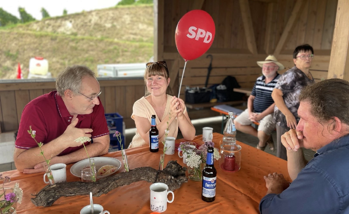 Drei Menschen sitzen an einem Tisch. Die Frau in der Mitte hat einen roten SPD-Luftbalon.